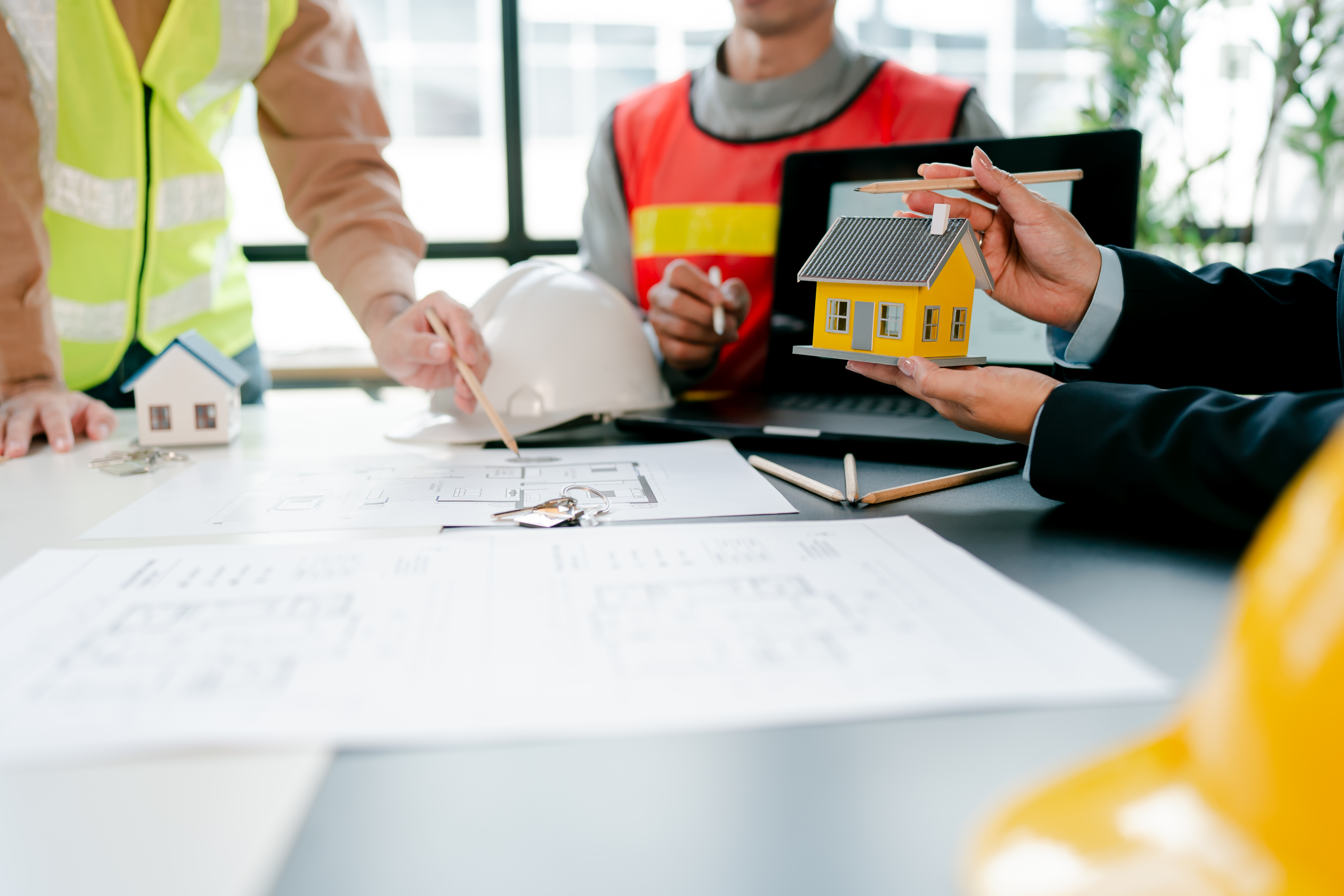 Business people collaborating on an architectural project, discussing house plans, construction, and property investment with building models, blueprints, and hard hats on a desk